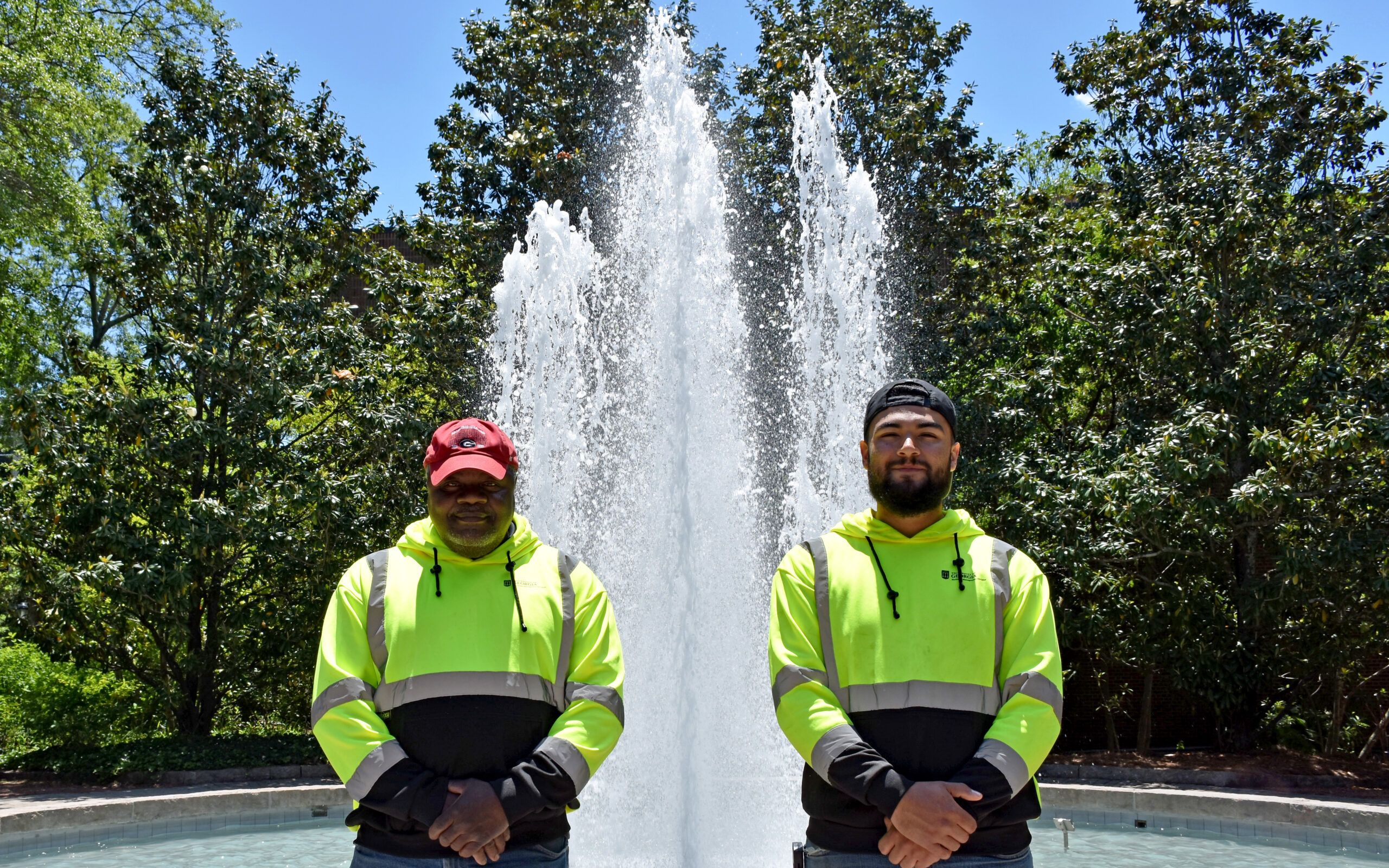 two men in yellow work shirts posing in front of fountain outside