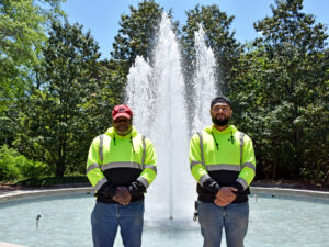 two men in yellow work shirts posing in front of fountain outside