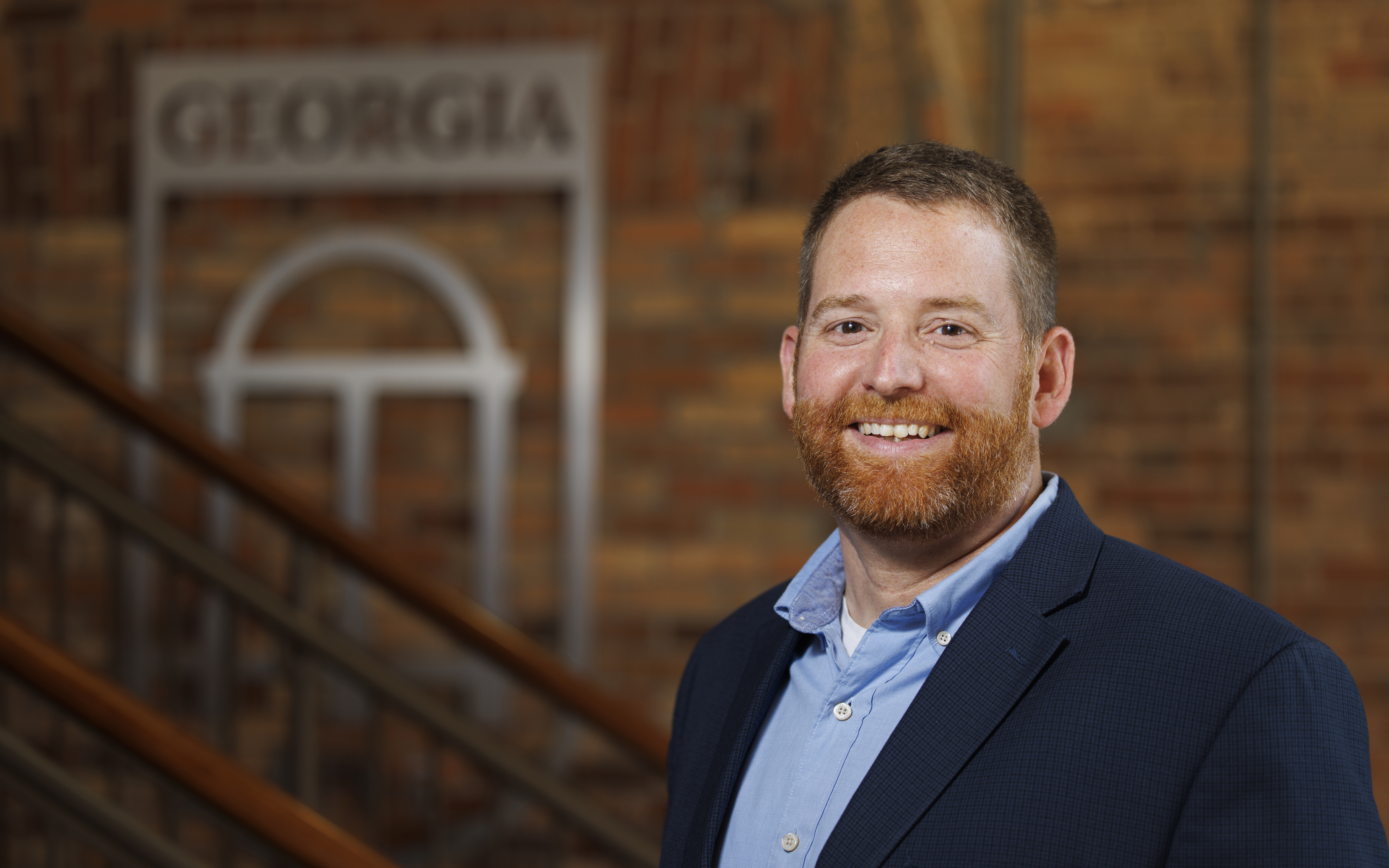 A smiling man in a blue button-down shirt and navy blazer stands in front of a brick wall and staircase, with a University of Georgia arch logo sign visible in the background.