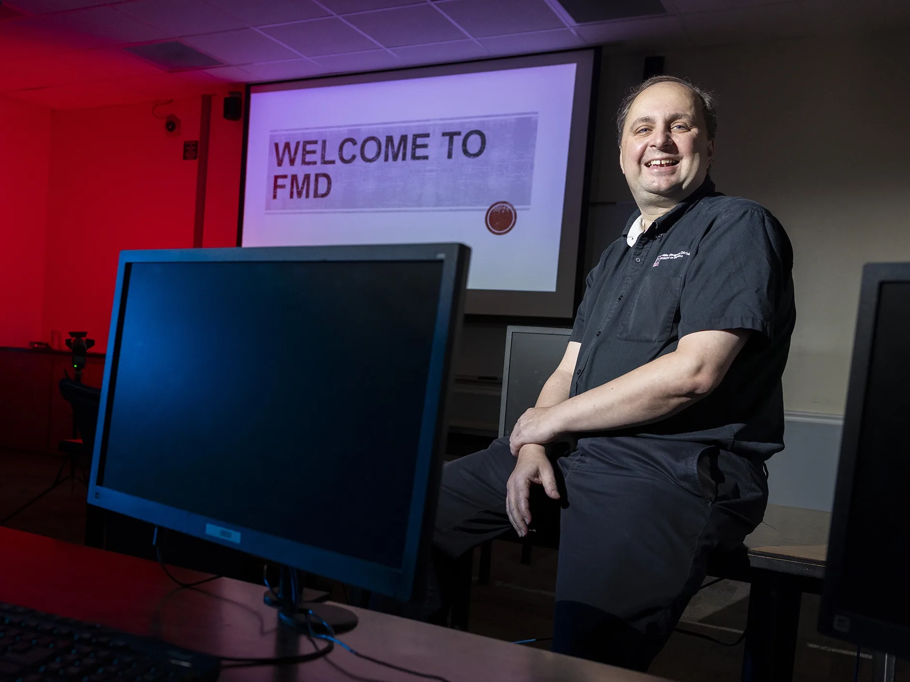 man in navy shirt and pants leaning against table with computer monitors around him and projected image in the back that reads "Welcome to FMD"